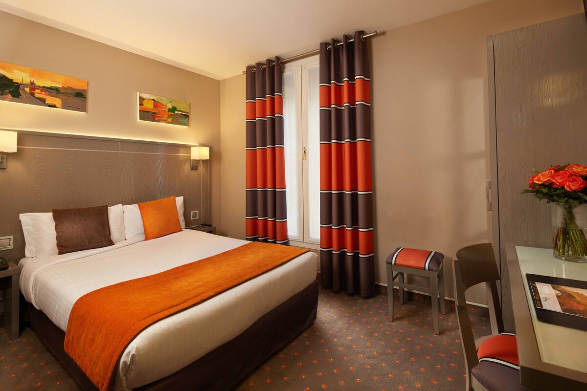 Een hotelkamer met een groot bed, een bureau met stoel, een kleine tafel met bloevas en twee schilderijen aan de muur.