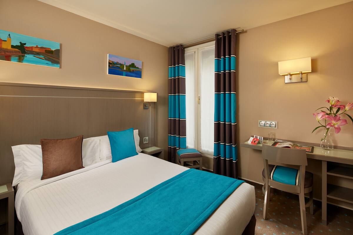 Een hotelkamer met een groot bed, een bureau met stoel, een klein tafeltje, een lamp en twee schilderijen aan de muur.