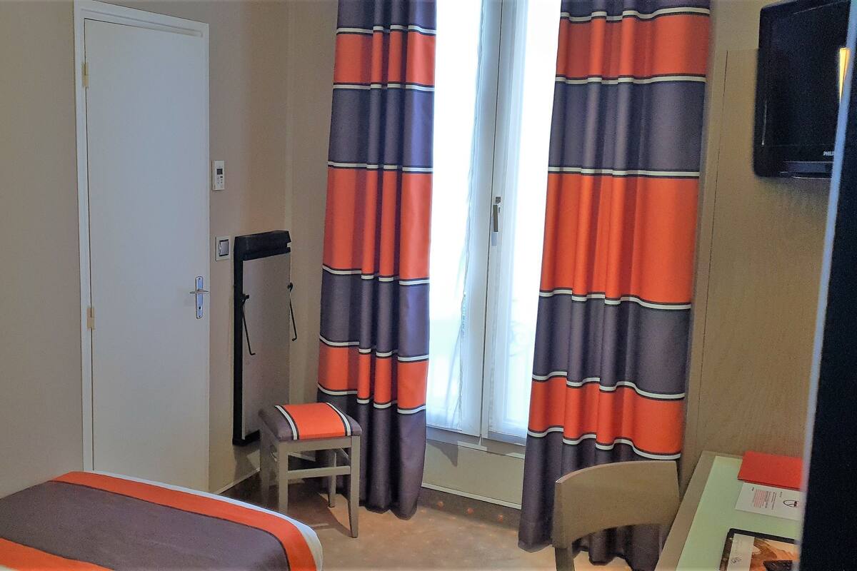 Een hotelkamer met een bed, een stoel, een bureau, een lamp, gordijnen en een schilderij aan de muur.