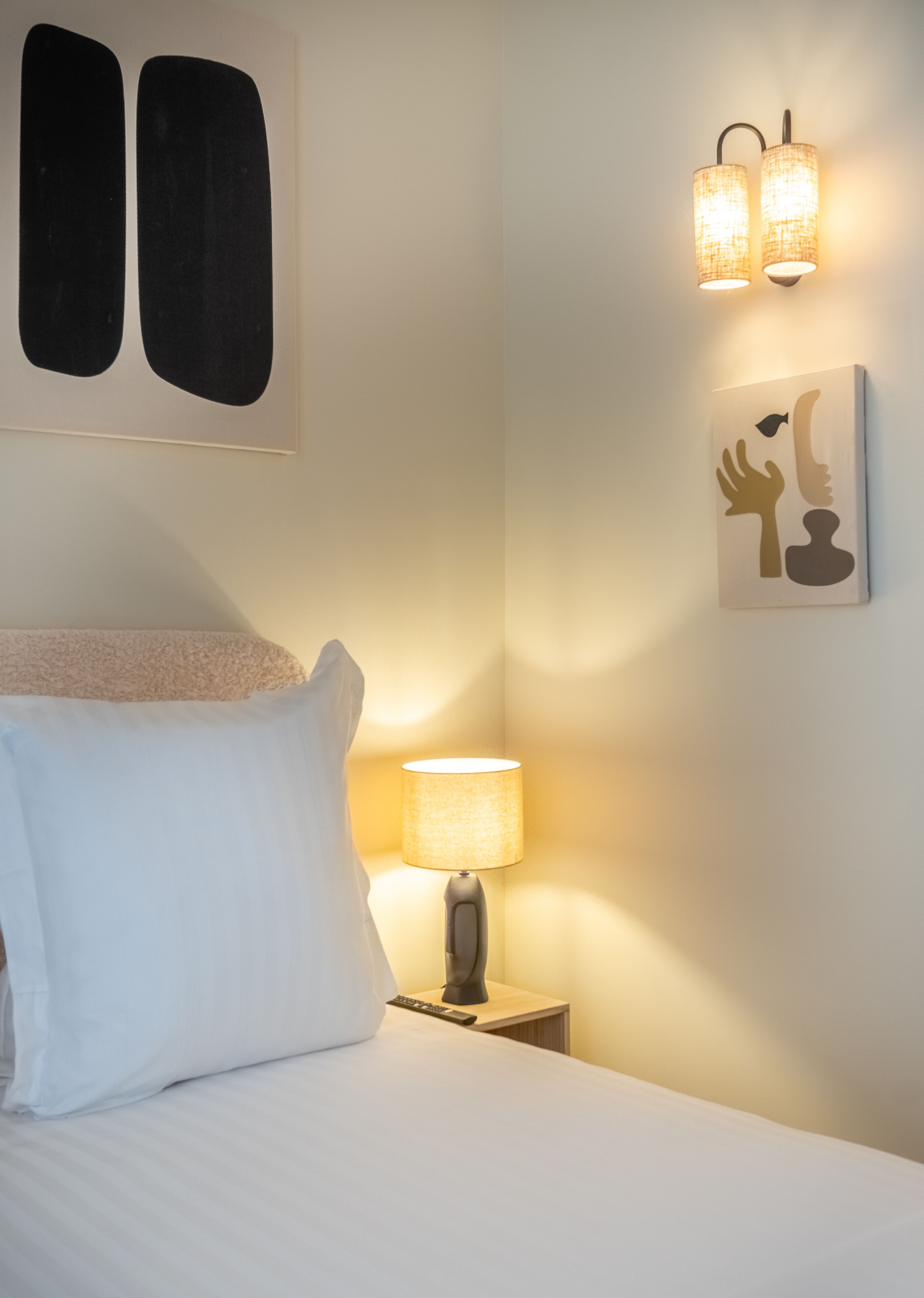 Un lit bien fait, avec une literie blanche, une lampe de chevet et une œuvre d’art accrochée au mur.