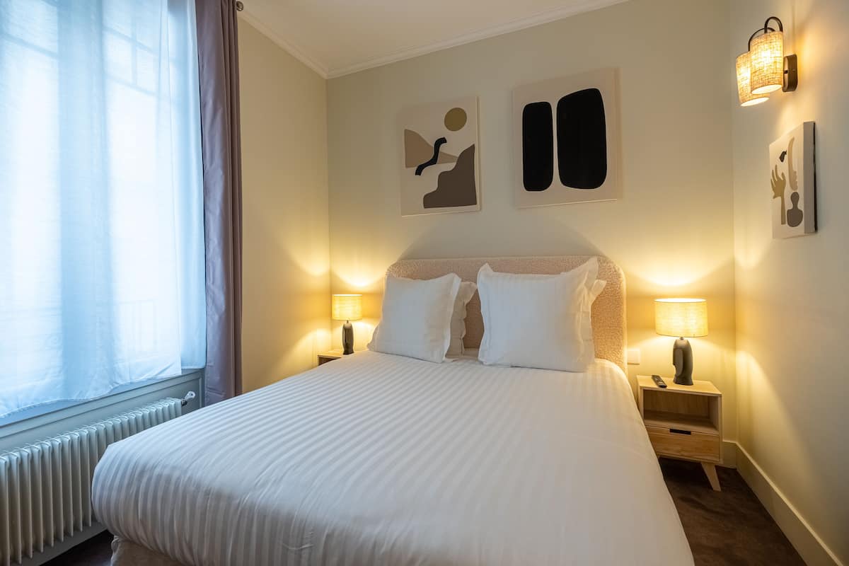 Uma cama bem arrumada com lençóis brancos, duas luminárias de cabeceira e arte abstrata na parede.