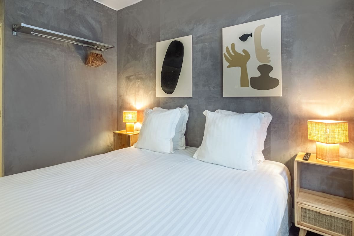 Cama bem arrumada com lençóis brancos, duas luminárias de cabeceira e arte abstrata na parede.