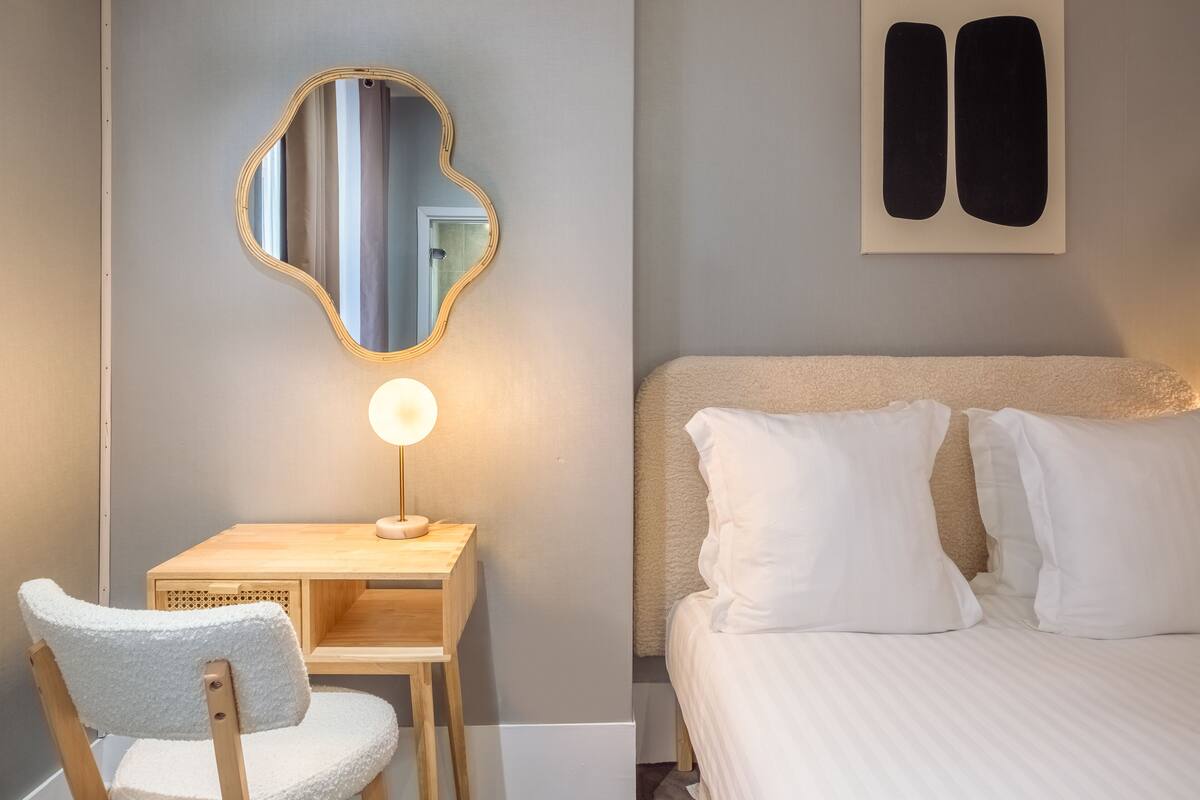 Um quarto com uma cama, uma cadeira, uma escrivaninha com luminária e um espelho na parede.