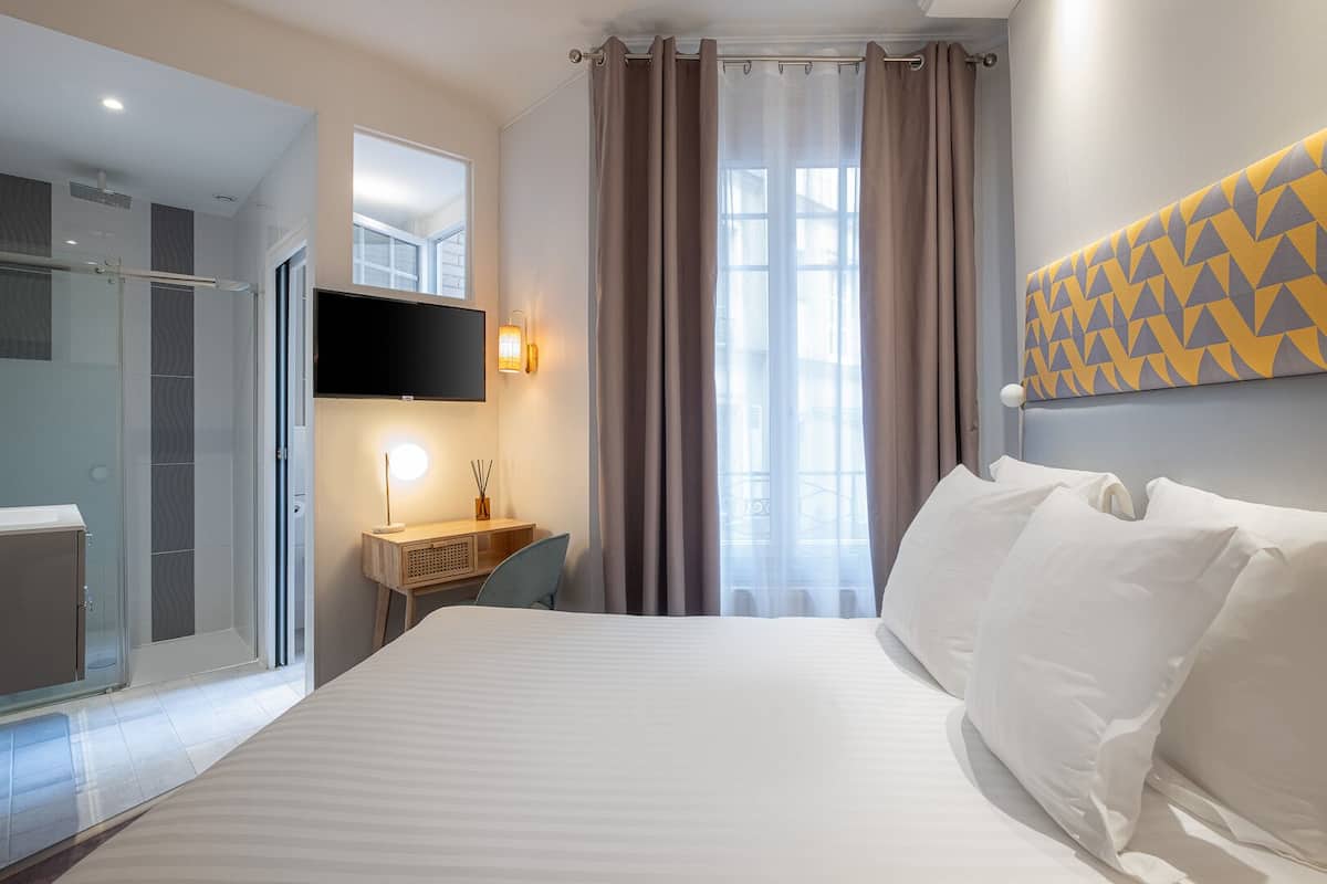 Quarto de hotel com cama, escrivaninha com luminária, televisão e área de chuveiro.