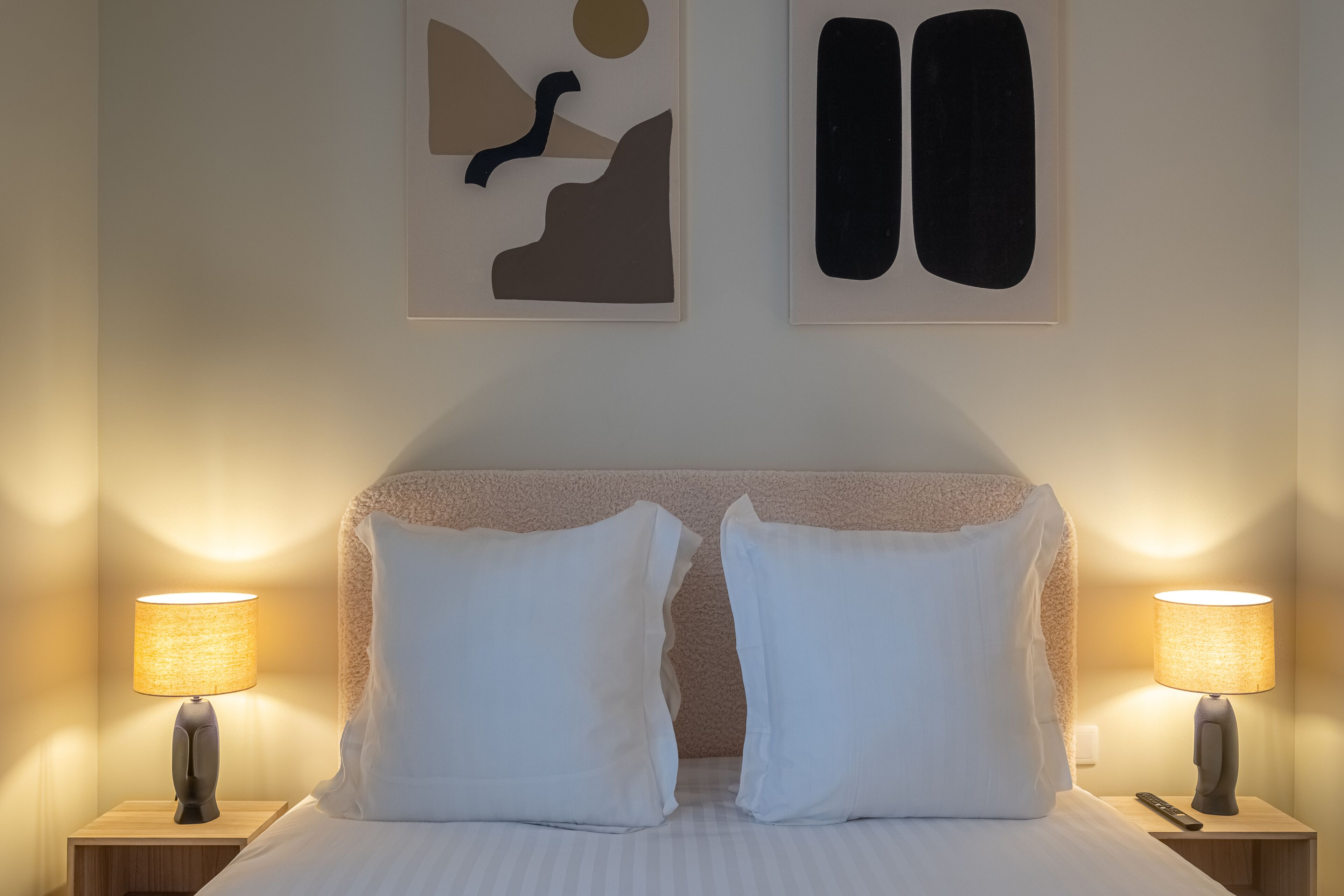 Un letto ben rifatto con lenzuola bianche e due cuscini, affiancato da due comodini con lampade, di fronte a una parete decorata con opere d'arte astratte.