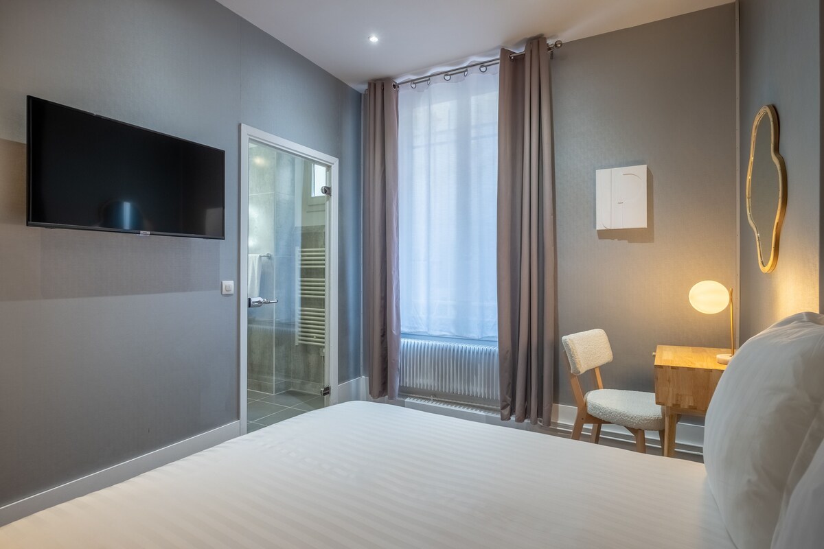 A hotel room with a bed, a TV, a desk with a chair, a window with curtains, and a radiator.