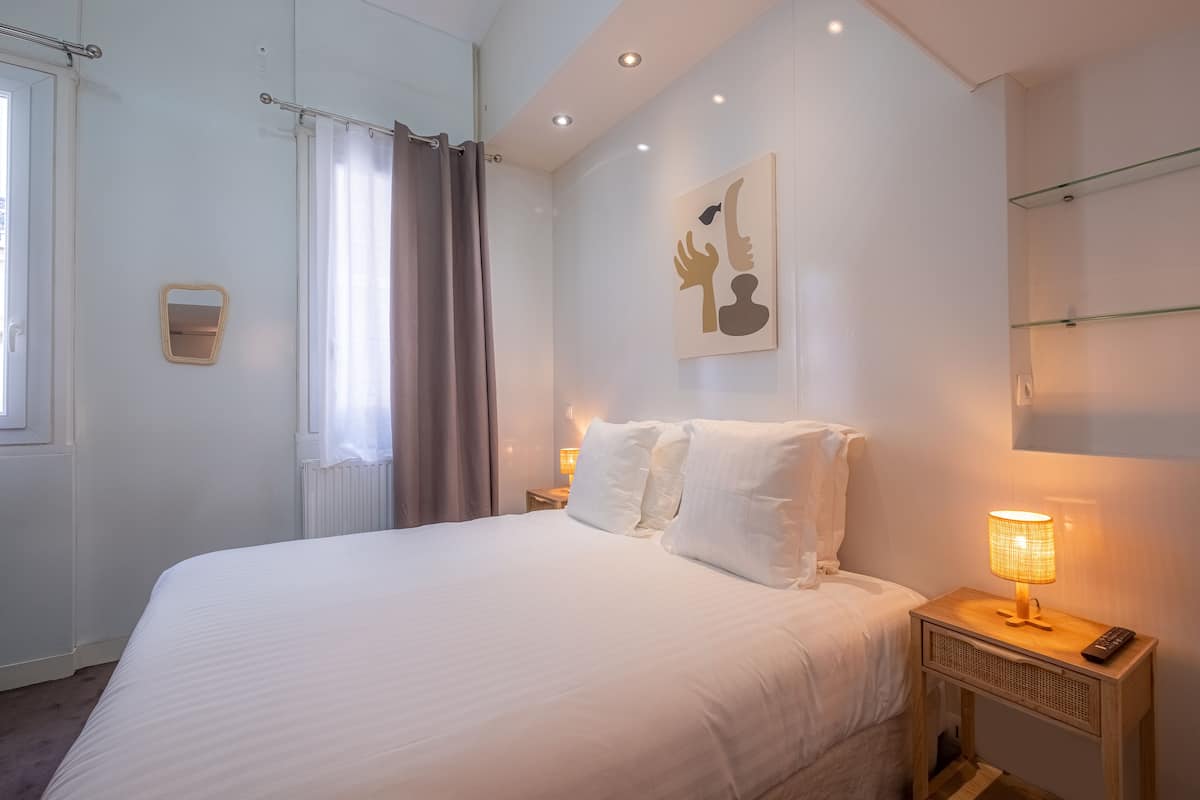 Cama bem arrumada com lençóis brancos, abajur e pequena mesa de cabeceira com controle remoto.