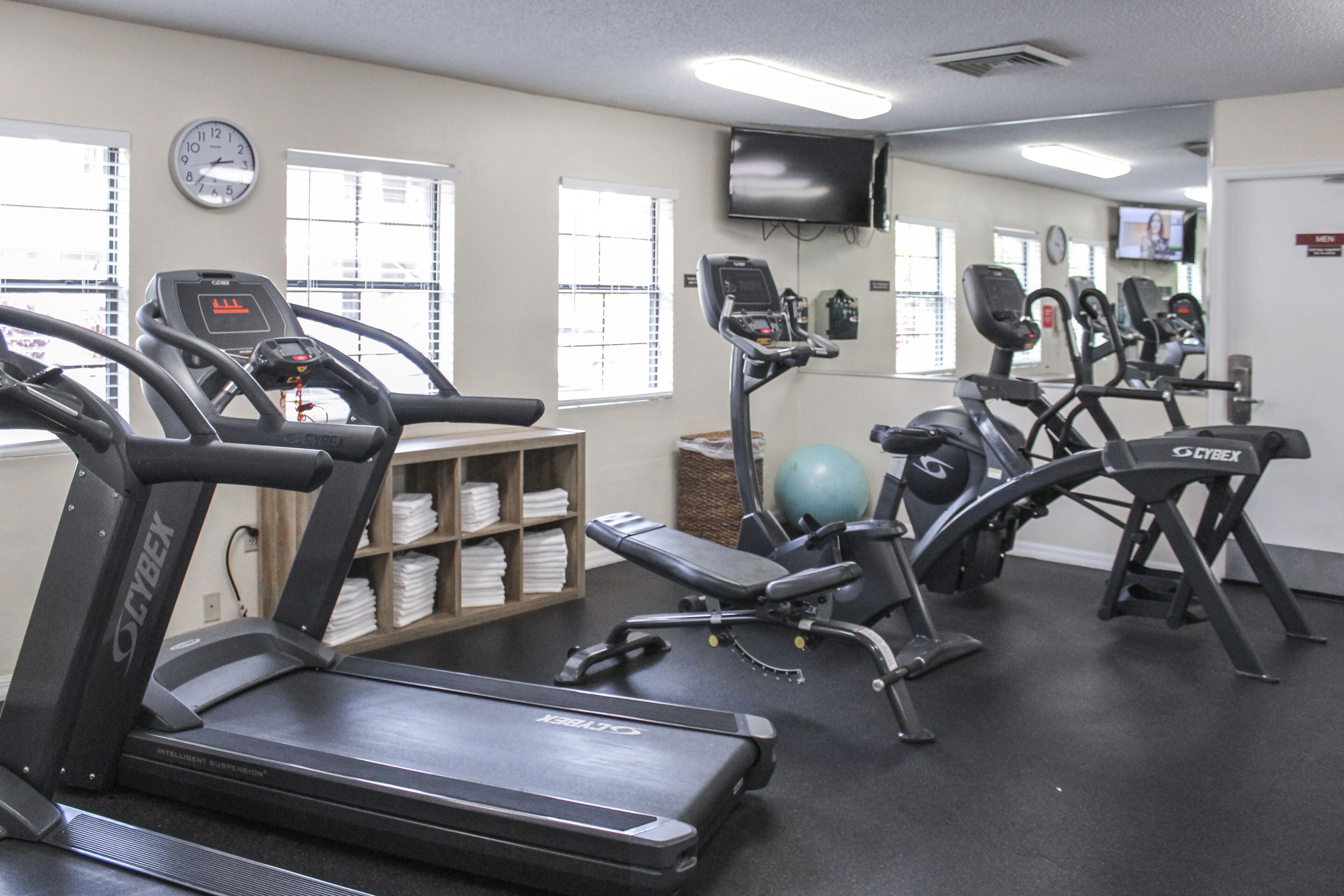 A gym with treadmills, elliptical machines, and a stationary bike. There are windows with blinds, a clock, and a television on the wall.