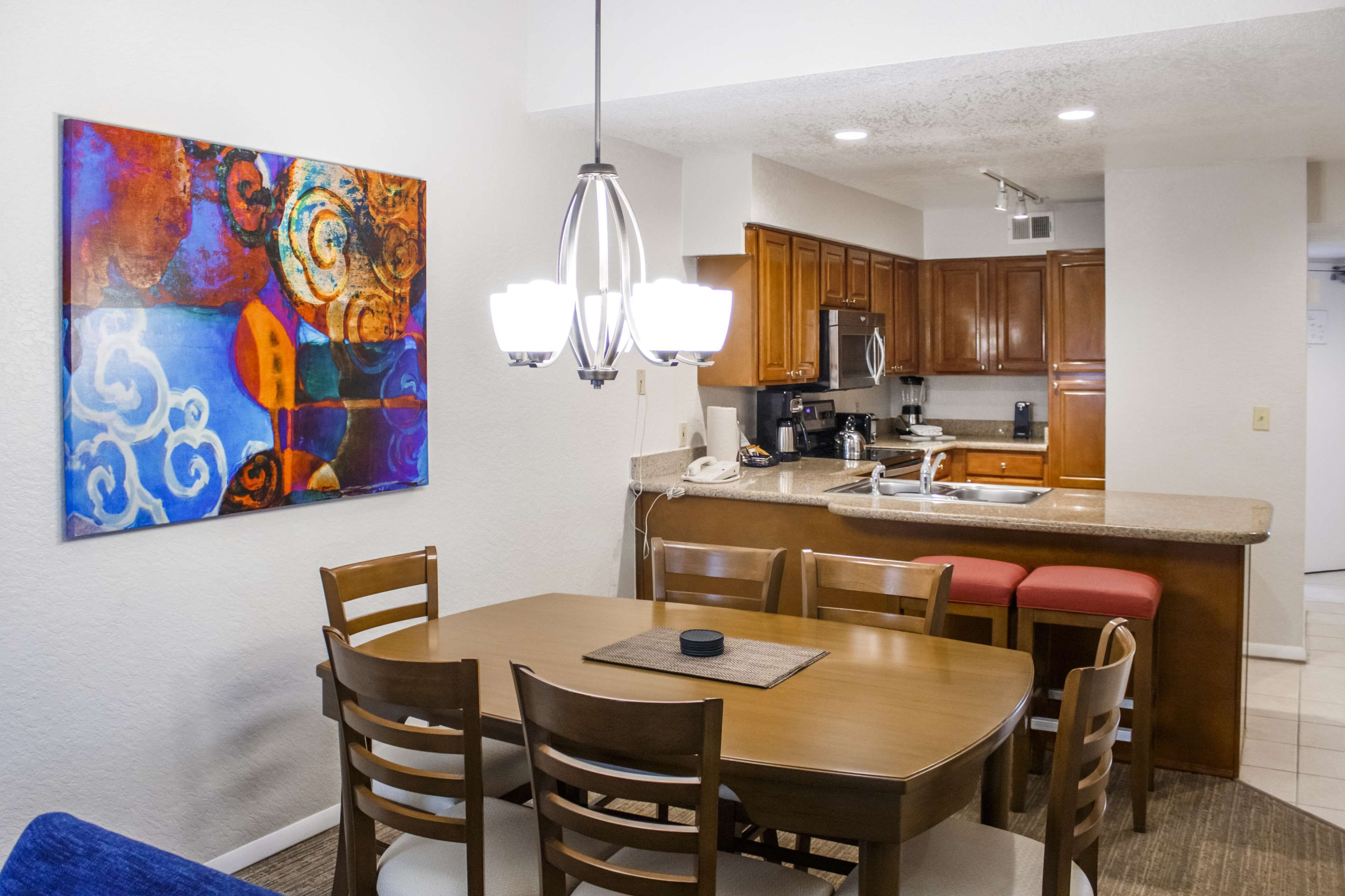 A dining area with a wooden table and chairs, a kitchen in the background, and a colorful abstract painting on the wall.