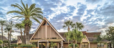 A hotel with a prominent triangular roof, surrounded by palm trees and a stone wall with orange flowers.