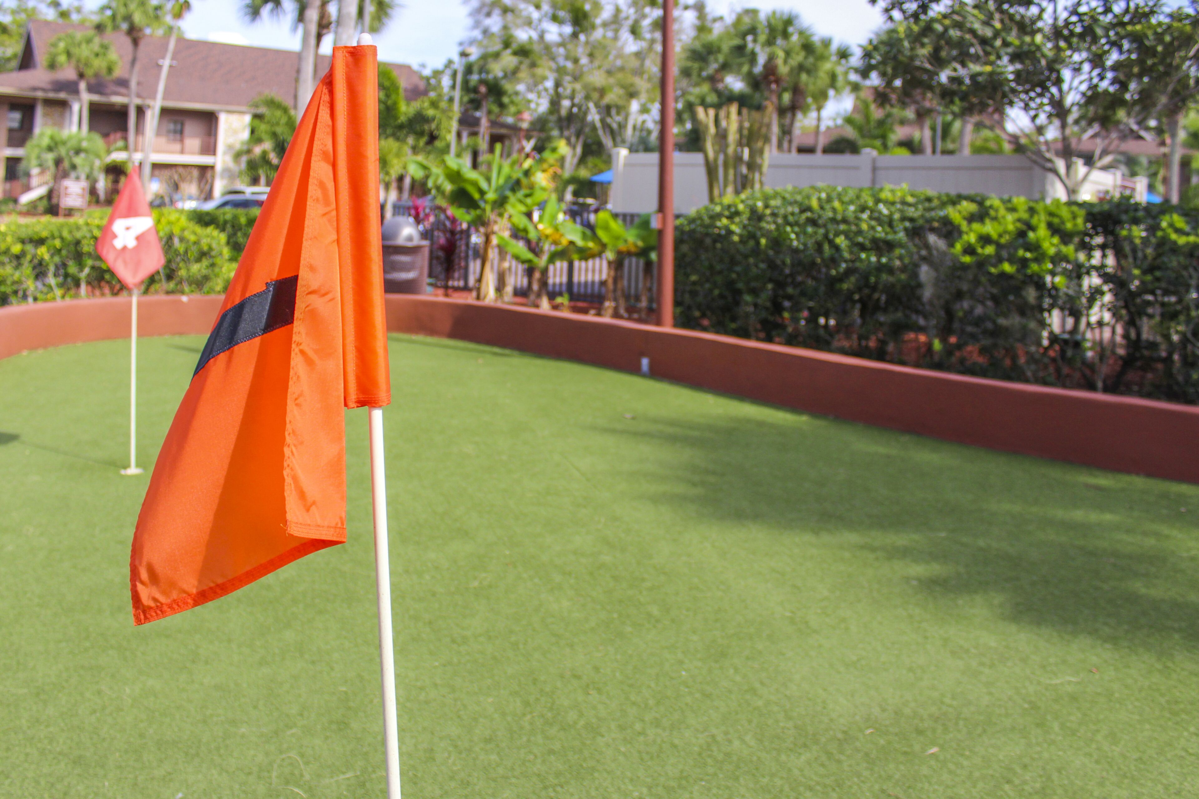 A golf flag on a green with a white pole, surrounded by a red border and a hedge.