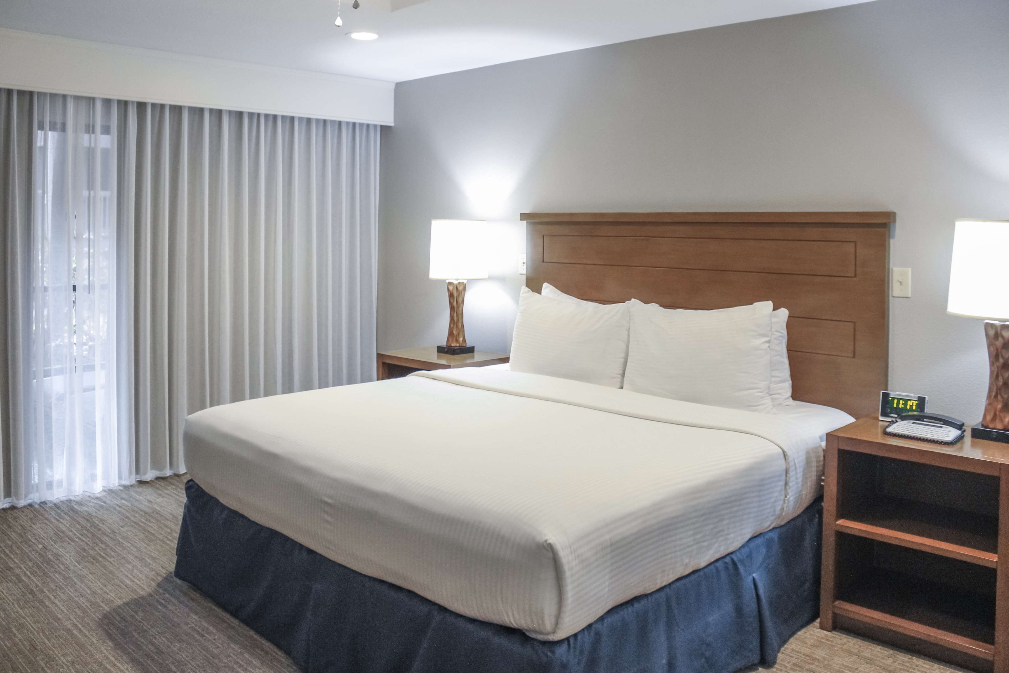 Une chambre d’hôtel avec un grand lit, deux tables de chevet, une petite table, une lampe et une fenêtre avec des rideaux.