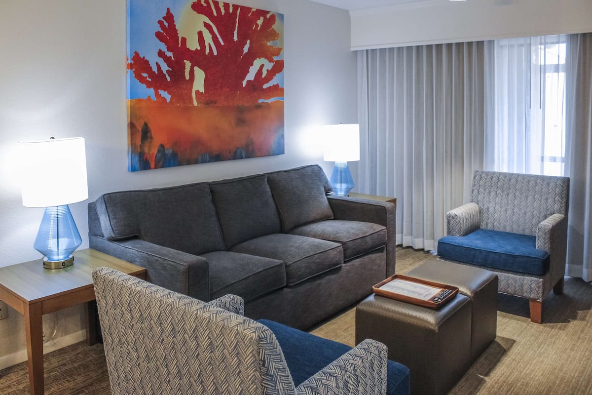 A modern hotel room with a grey sofa, patterned armchair, and ottoman, a blue lamp, and a large abstract painting on the wall.