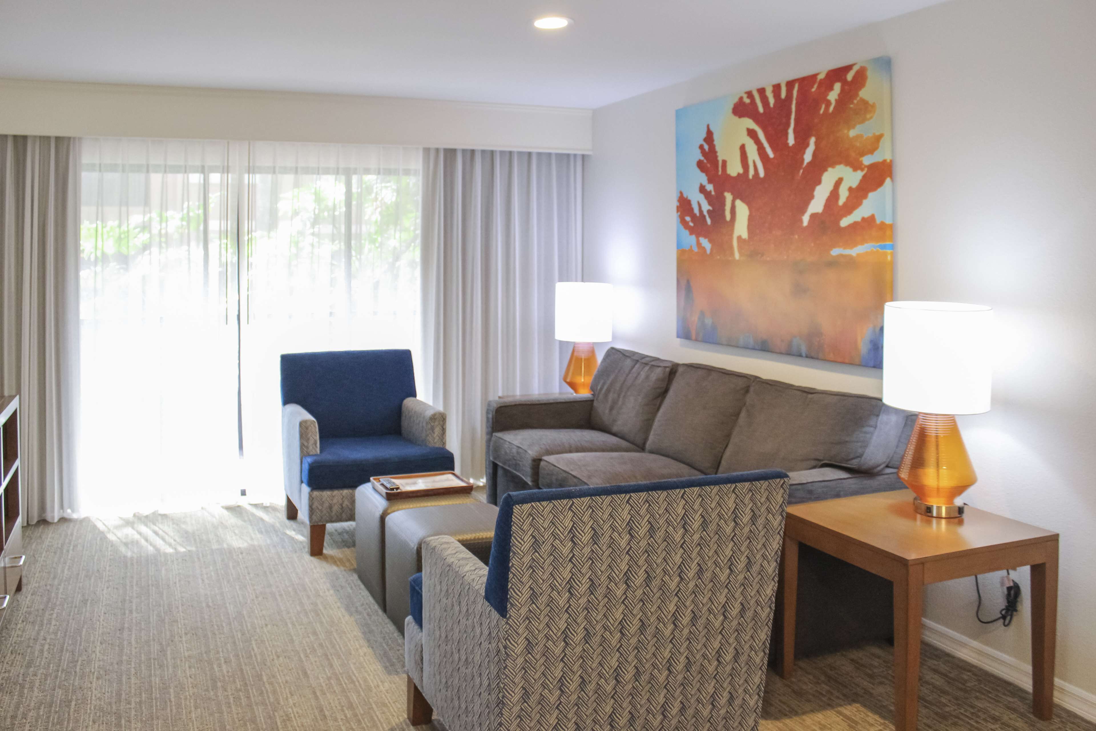 Habitación de hotel con sofá, sillón y una pequeña mesa con lámpara. Cuenta con un ventanal con cortinas y un cuadro en la pared.