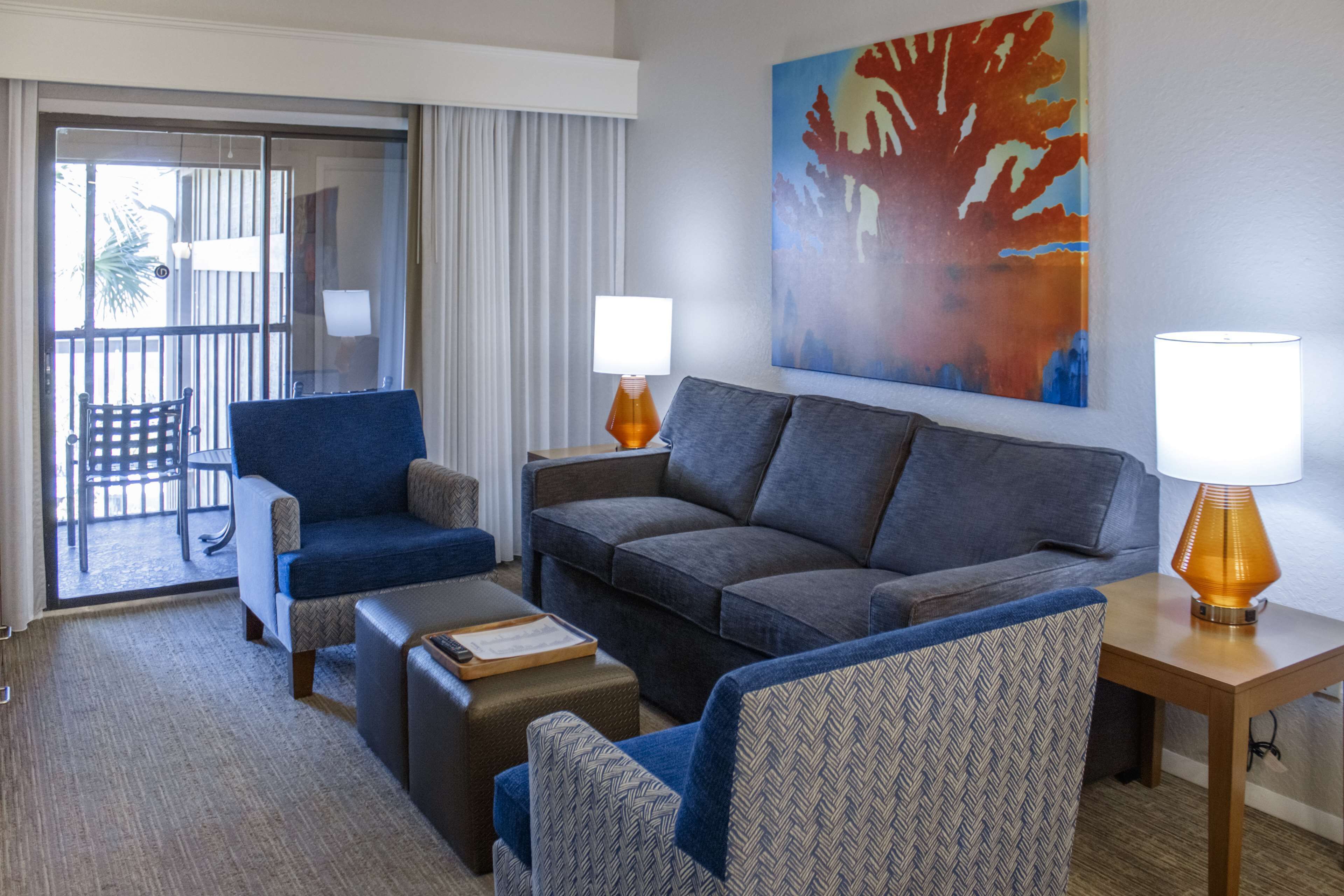 A modern hotel room with a grey sofa, blue armchair, and patterned armchair. There is a wooden side table with a lamp, a small ottoman with a tray, and a large window with curtains.