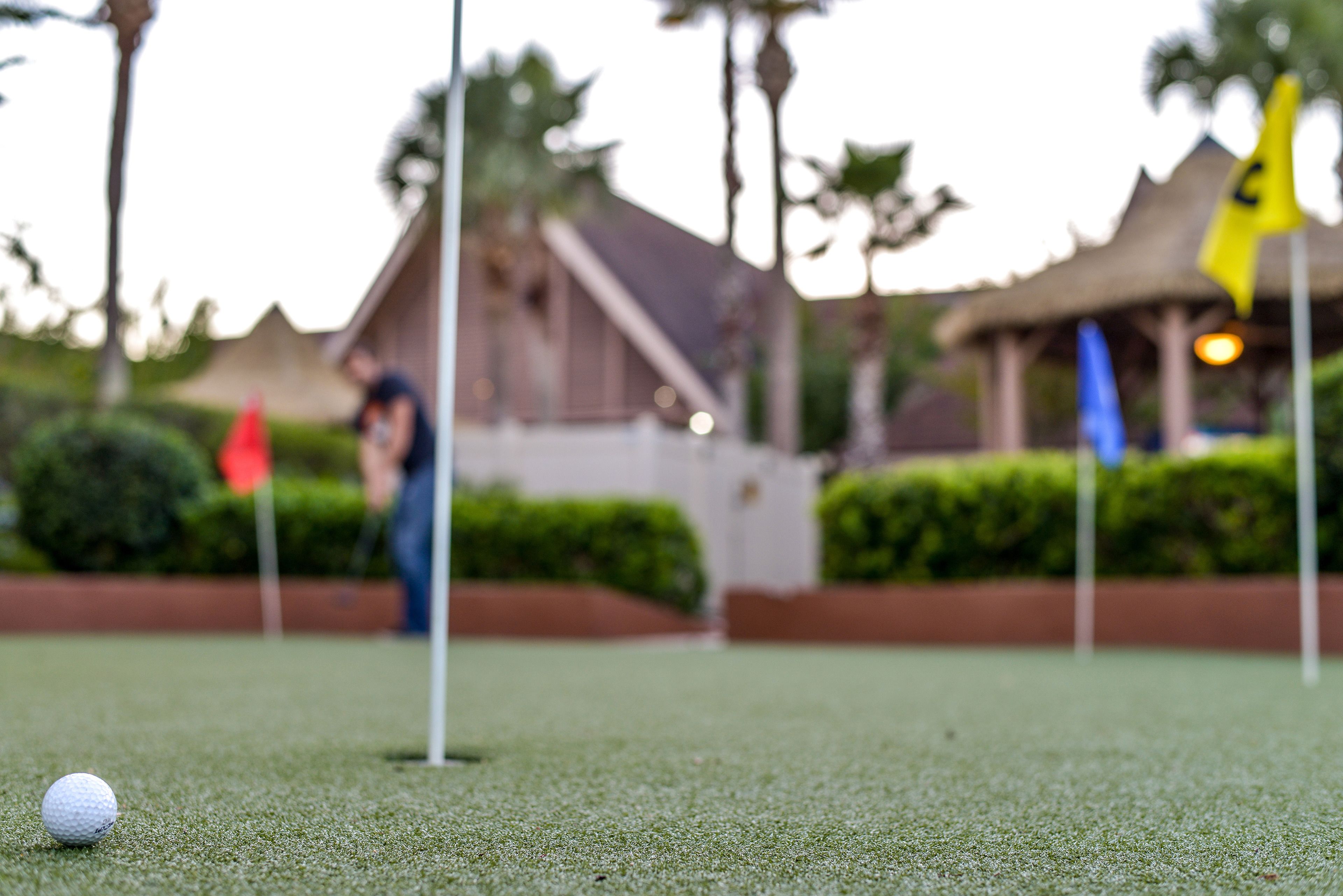 Une balle de golf sur un green, avec des drapeaux et un bâtiment en arrière-plan.