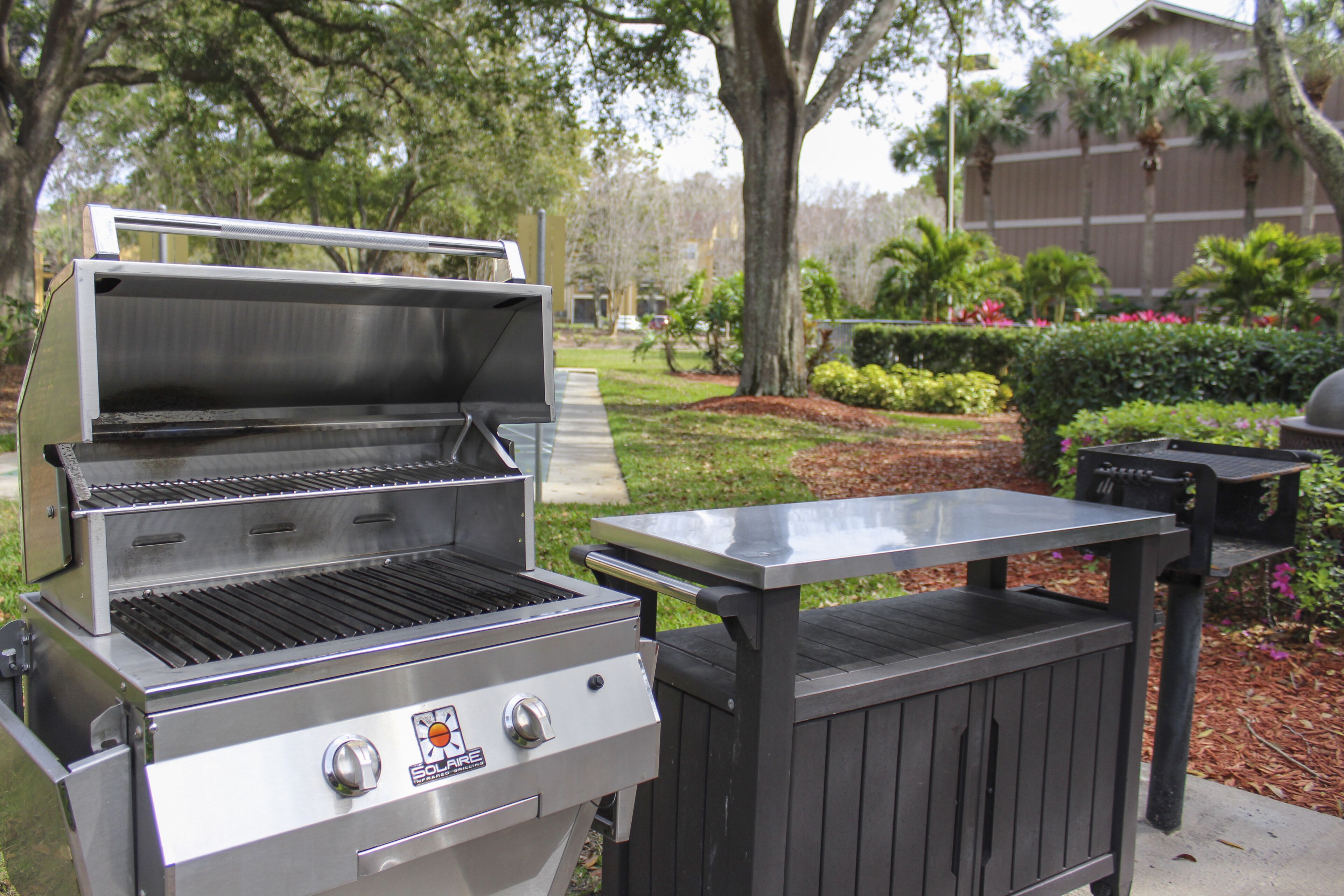 Un barbecue en acier inoxydable doté d’une table d’appoint, installé dans un espace extérieur aménagé avec des arbres et des arbustes.