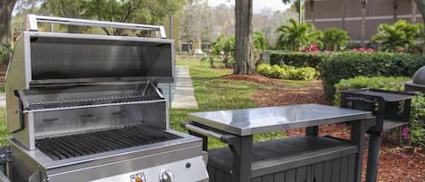 A stainless steel grill with a side table, set in a landscaped outdoor area with trees and shrubs.