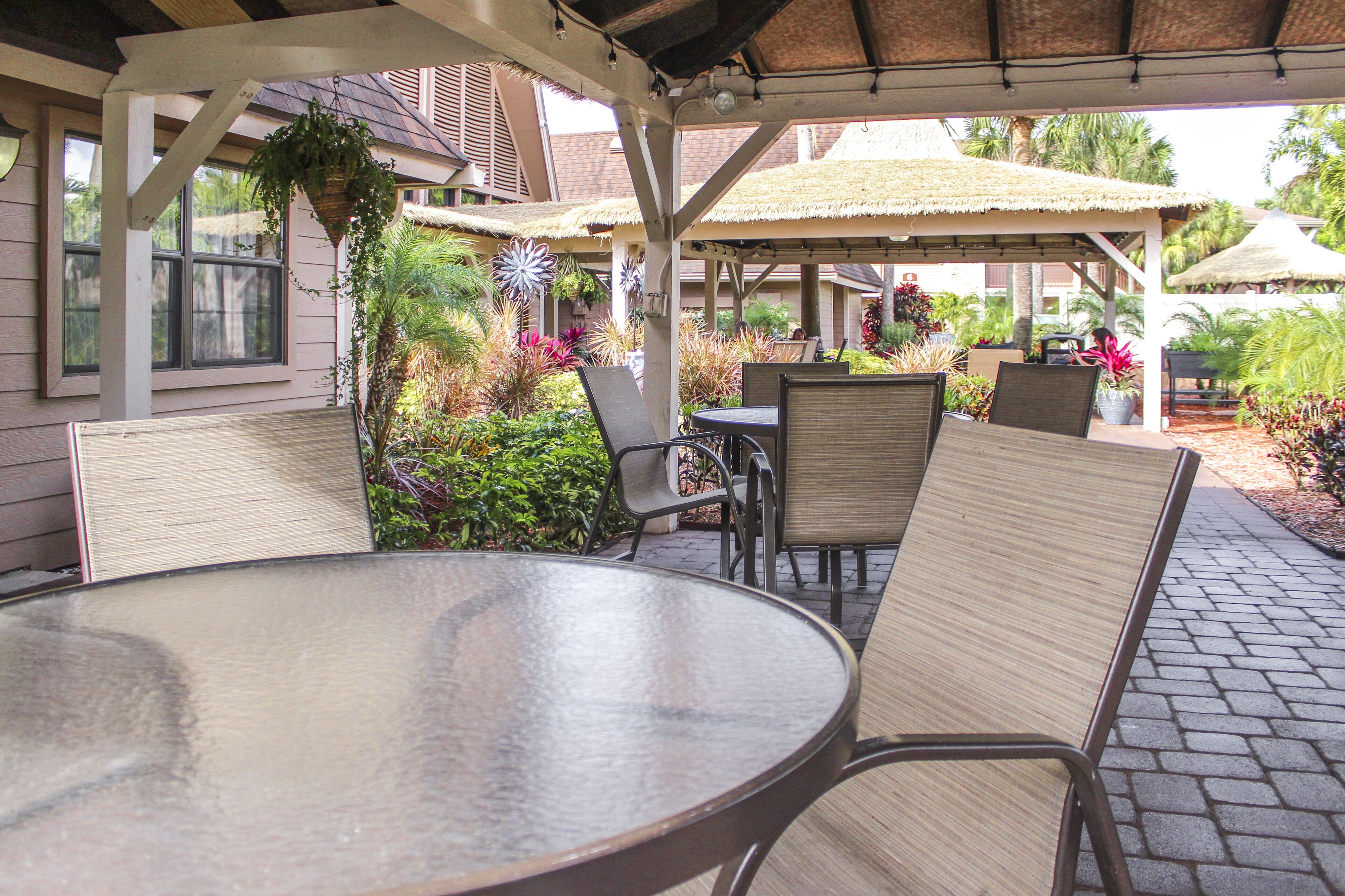 An outdoor patio with a round table and chairs, surrounded by potted plants and a thatched roof structure.
