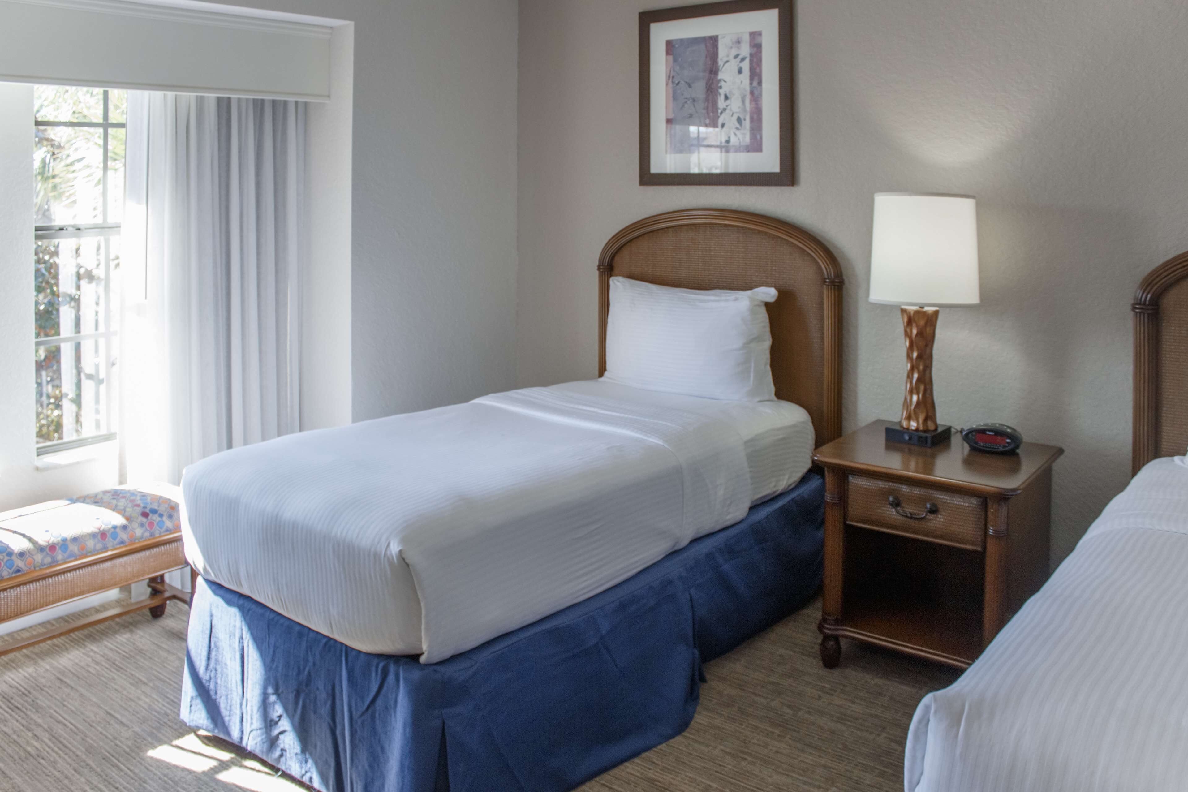 A hotel room with a single bed, a bedside table with a lamp, a small bedside table with a clock, and a window with curtains.