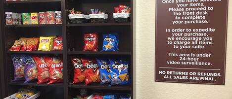 A pantry stocked with snacks and cereals, including Kellogg's Corn Flakes, Pop Weaver Butter, and Doritos. A sign instructs customers to charge items to their suite and notes 24-hour video surveillance.
