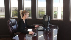 A person seated at a desk with a laptop, in a room with large windows and a dark wall. - The Greenwell Inn (Price)