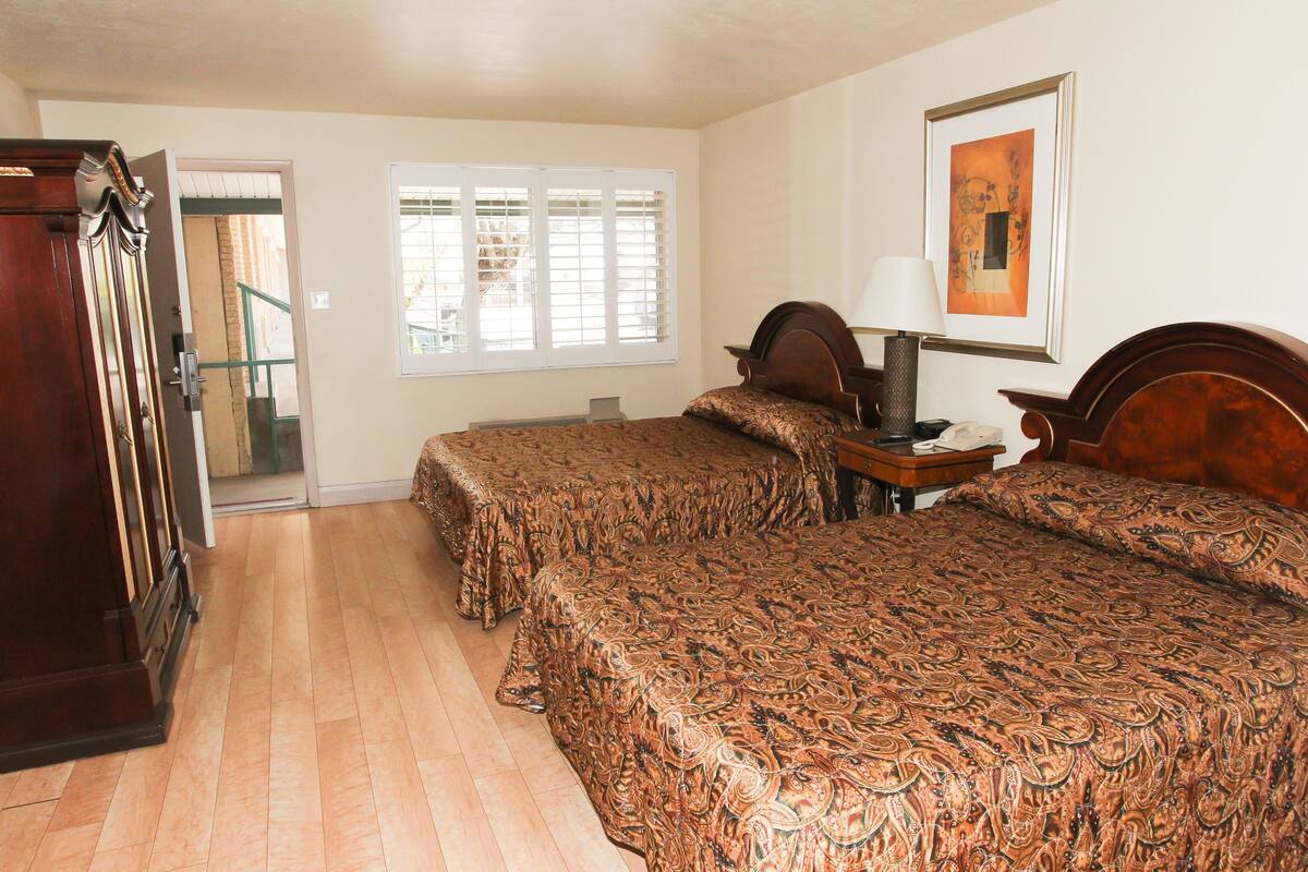 A hotel room with two beds, a wooden wardrobe, a desk with a lamp, and a window with blinds.