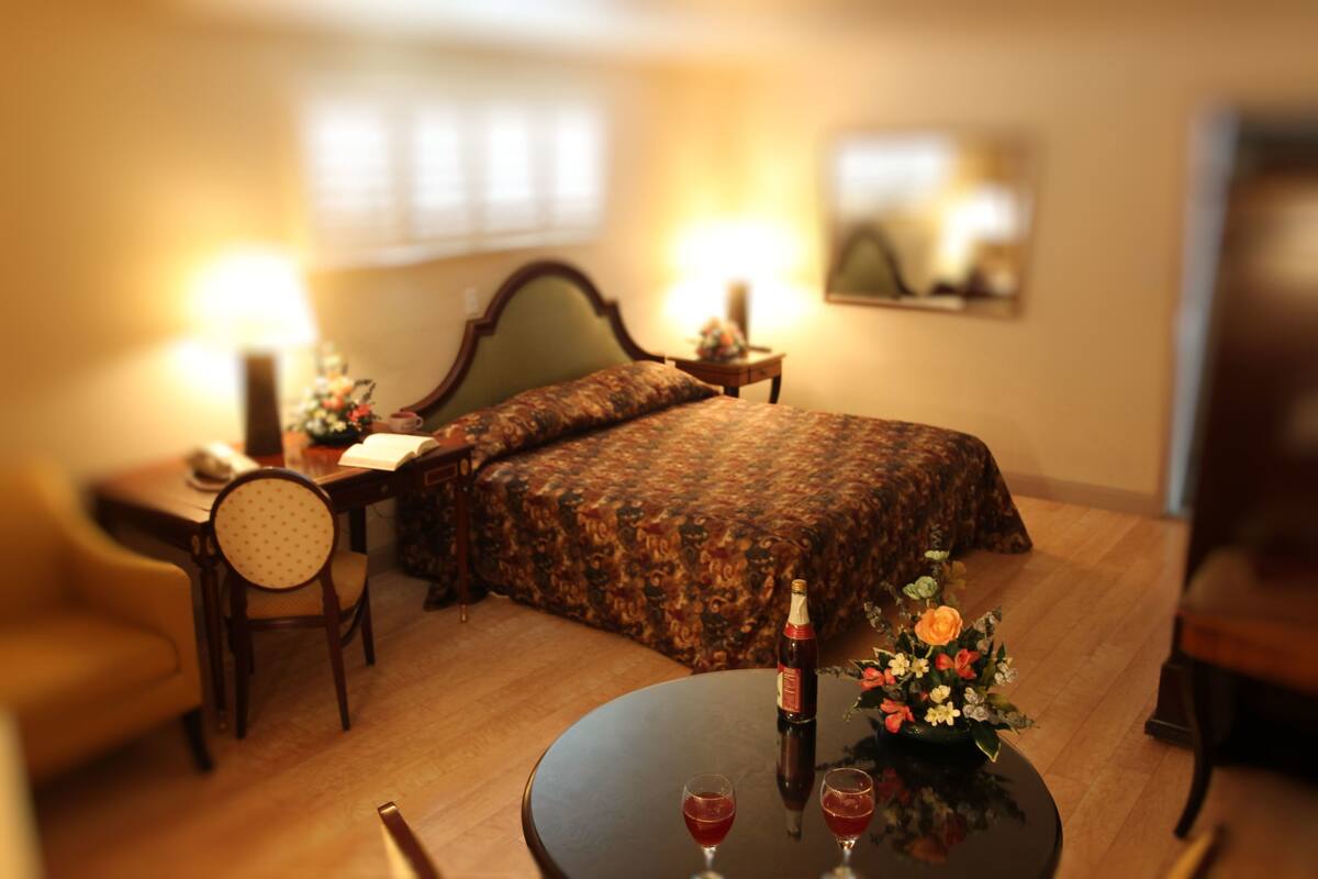A hotel room with a bed, a round table with two glasses, a bottle, and a flower arrangement.