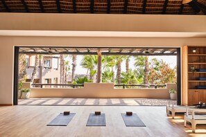 A spacious room with a large window overlooking palm trees and a building, wooden flooring, and exercise mats.
