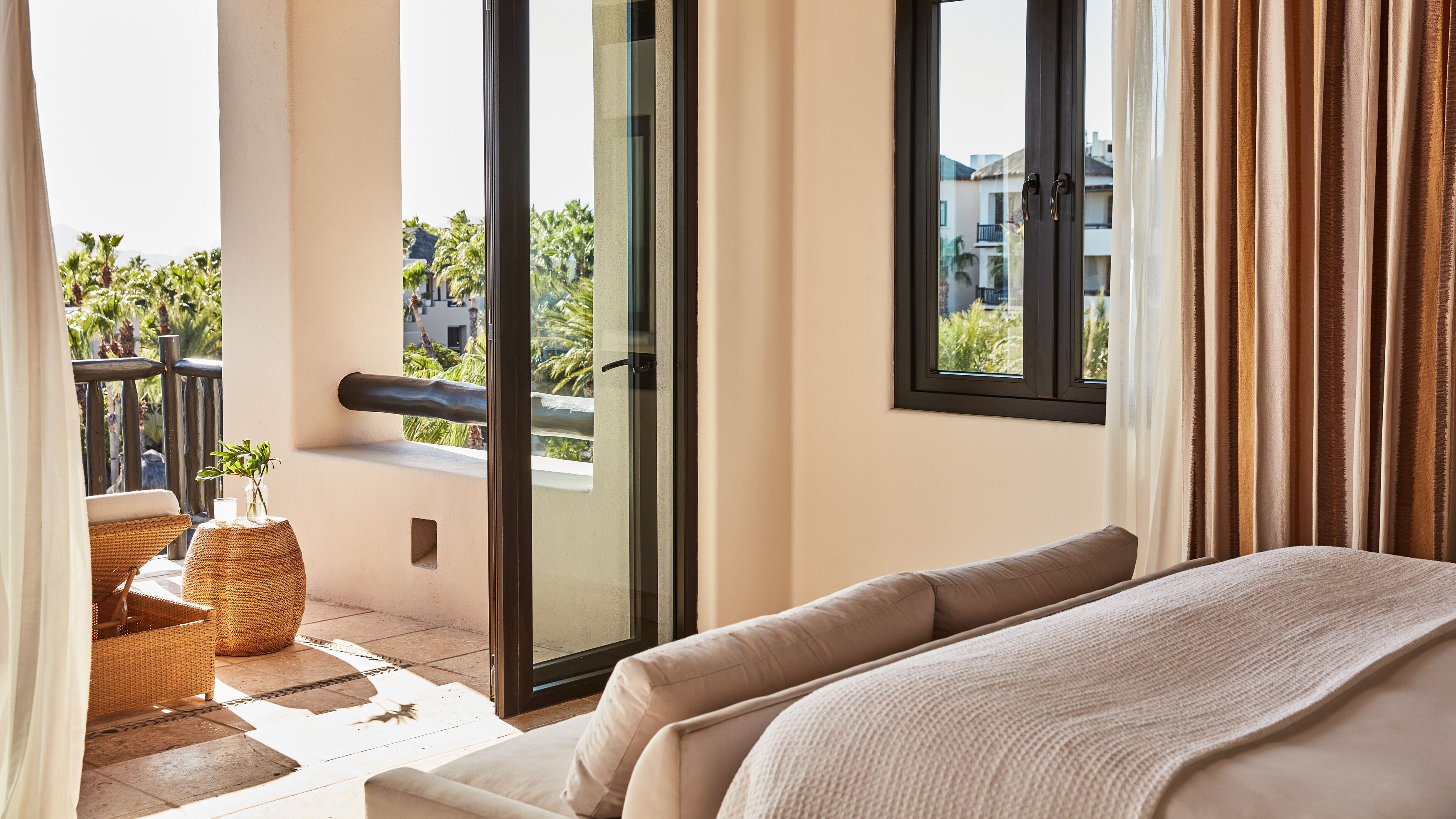 A hotel room with a balcony, a sofa, a small table with a plant, and a view of palm trees and buildings.