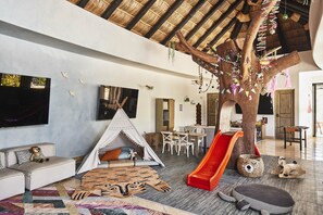 A spacious living area with a high ceiling, a large tree structure, a slide, and a cozy seating area with a sofa and cushions.