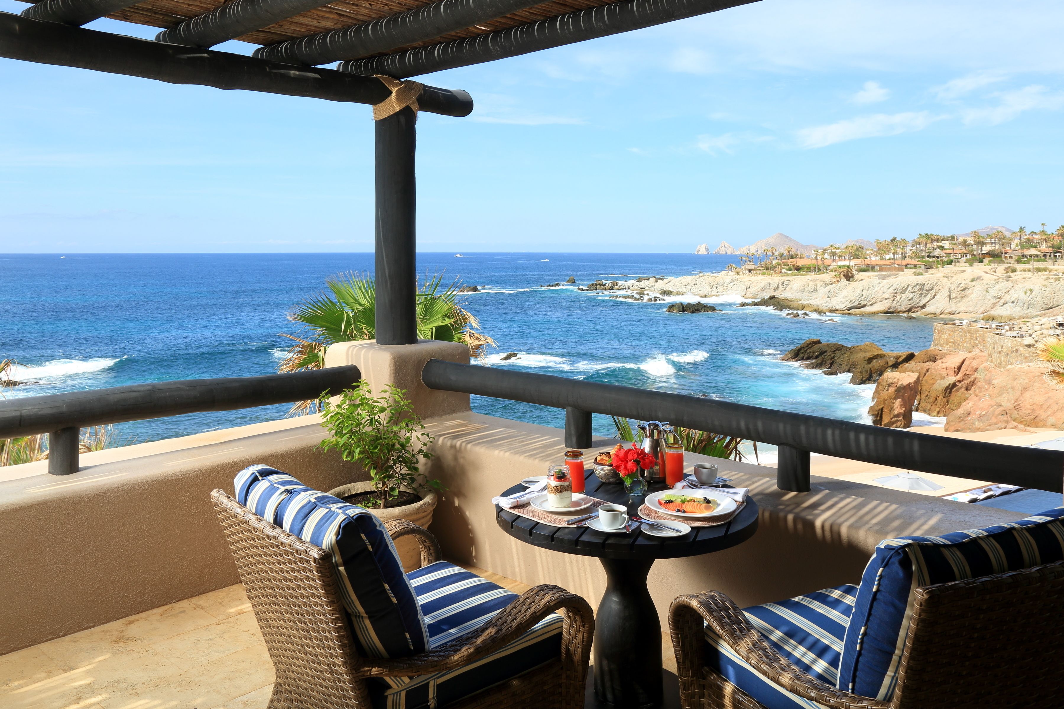 A balcony with wicker furniture, a table set with plates and drinks, and a view of the ocean and rocky coastline.