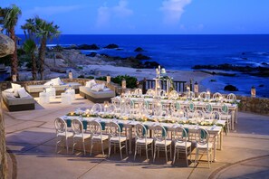 An outdoor dining area with tables and chairs set up for a meal, overlooking a beach with the ocean in the background.