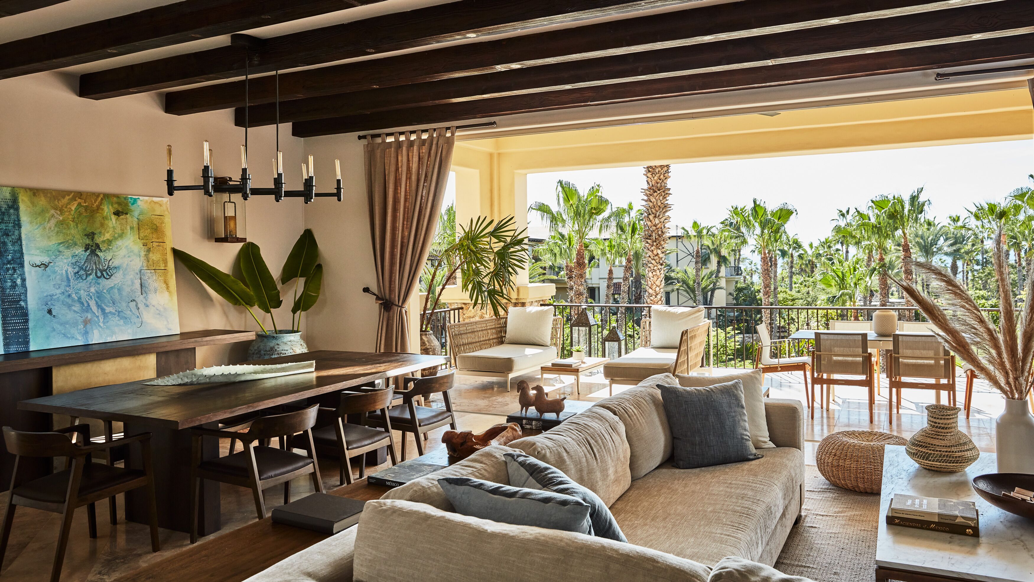 A spacious living area with a large sofa, dining table, and chairs. There is a balcony with plants and a view of palm trees.