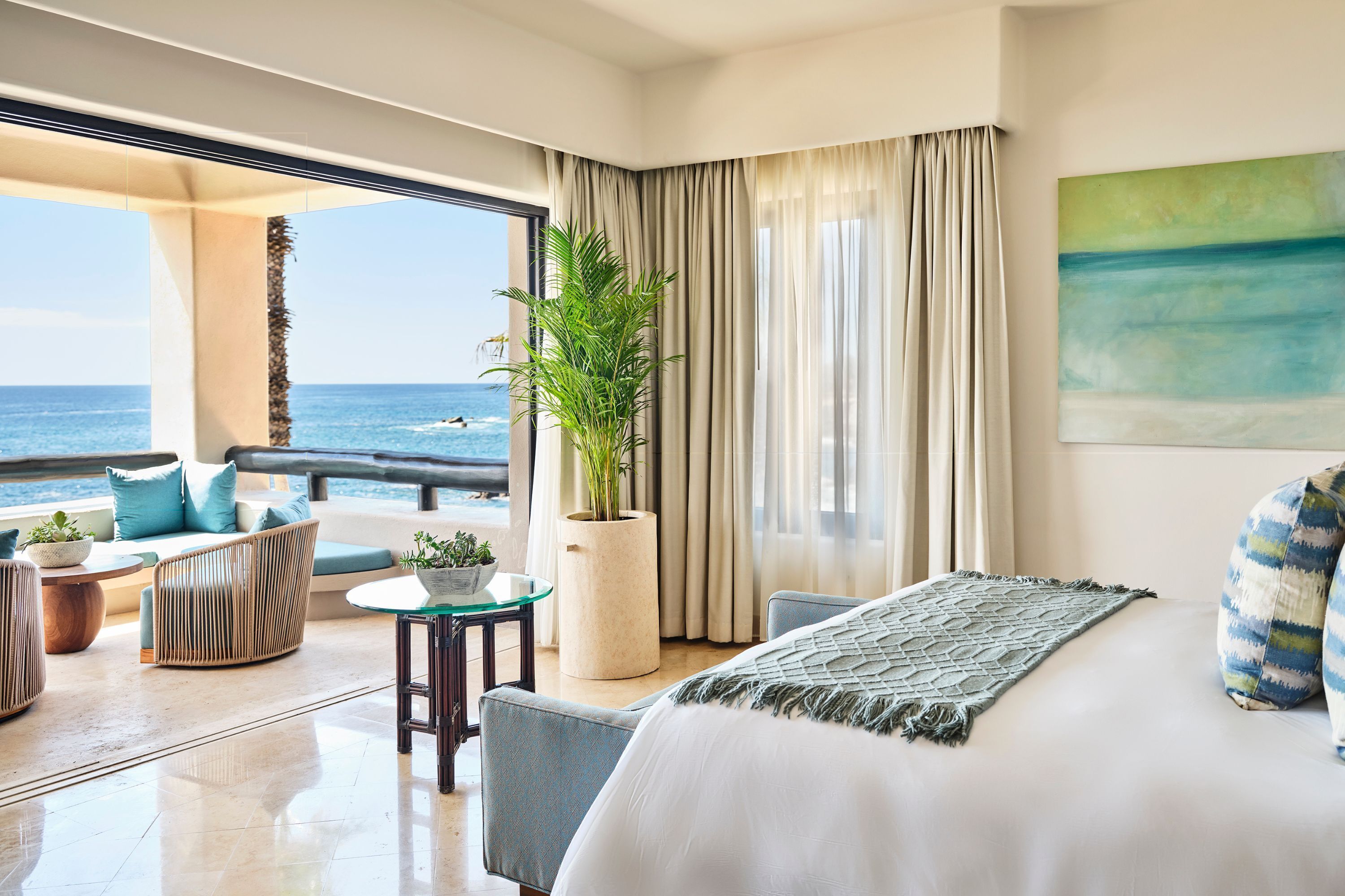 A hotel room with a balcony, a bed, a seating area with a plant, and a view of the ocean.