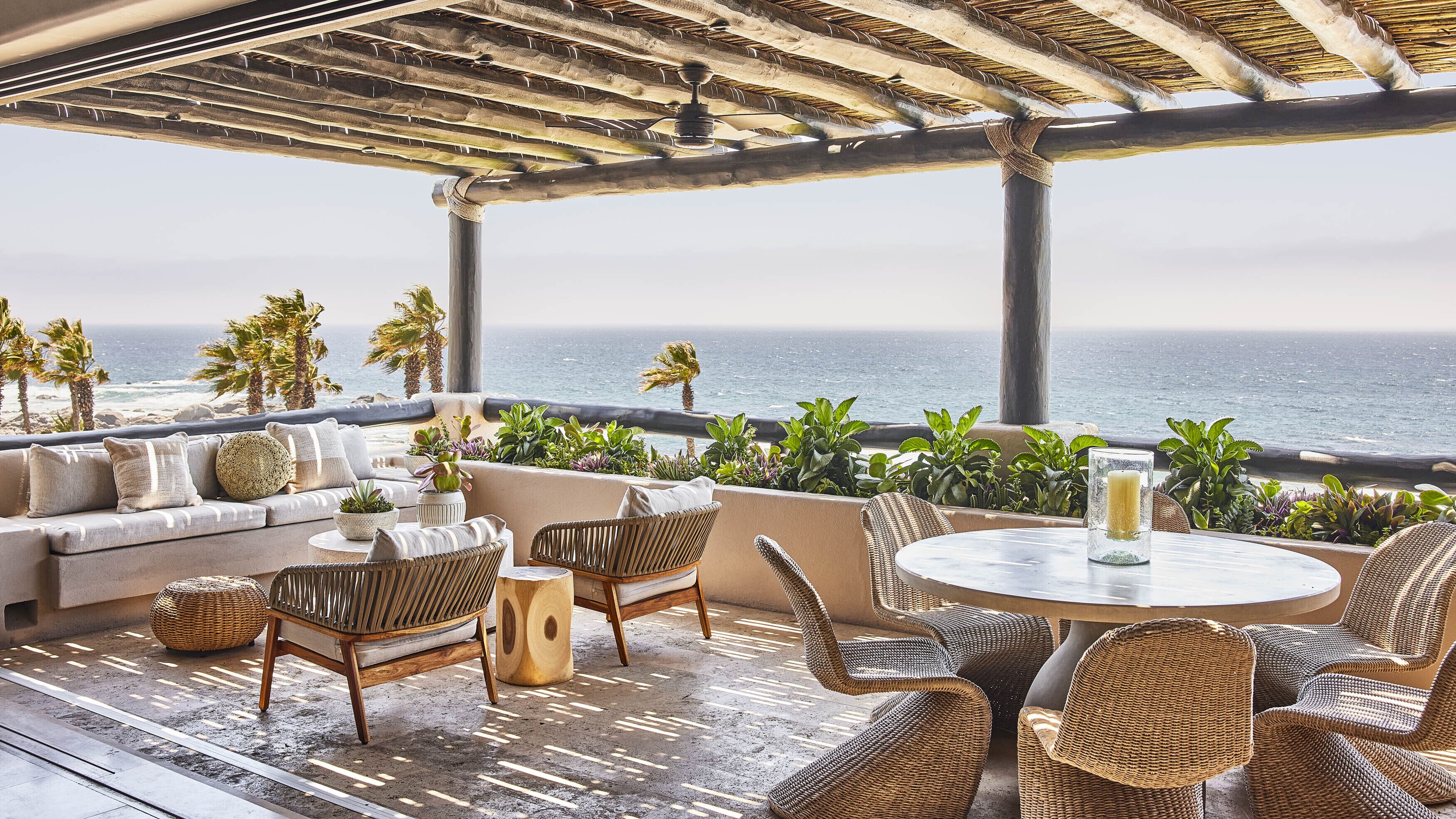 A coastal terrace with a sofa, wicker chairs, a round table, and potted plants.