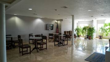 A spacious lobby with a high ceiling, recessed lighting, a flat-screen TV mounted on the wall, and several seating areas with chairs and tables.