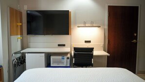 A hotel room with a bed, a desk with a flat-screen TV, a microwave, a small refrigerator, a towel rack, and a door.