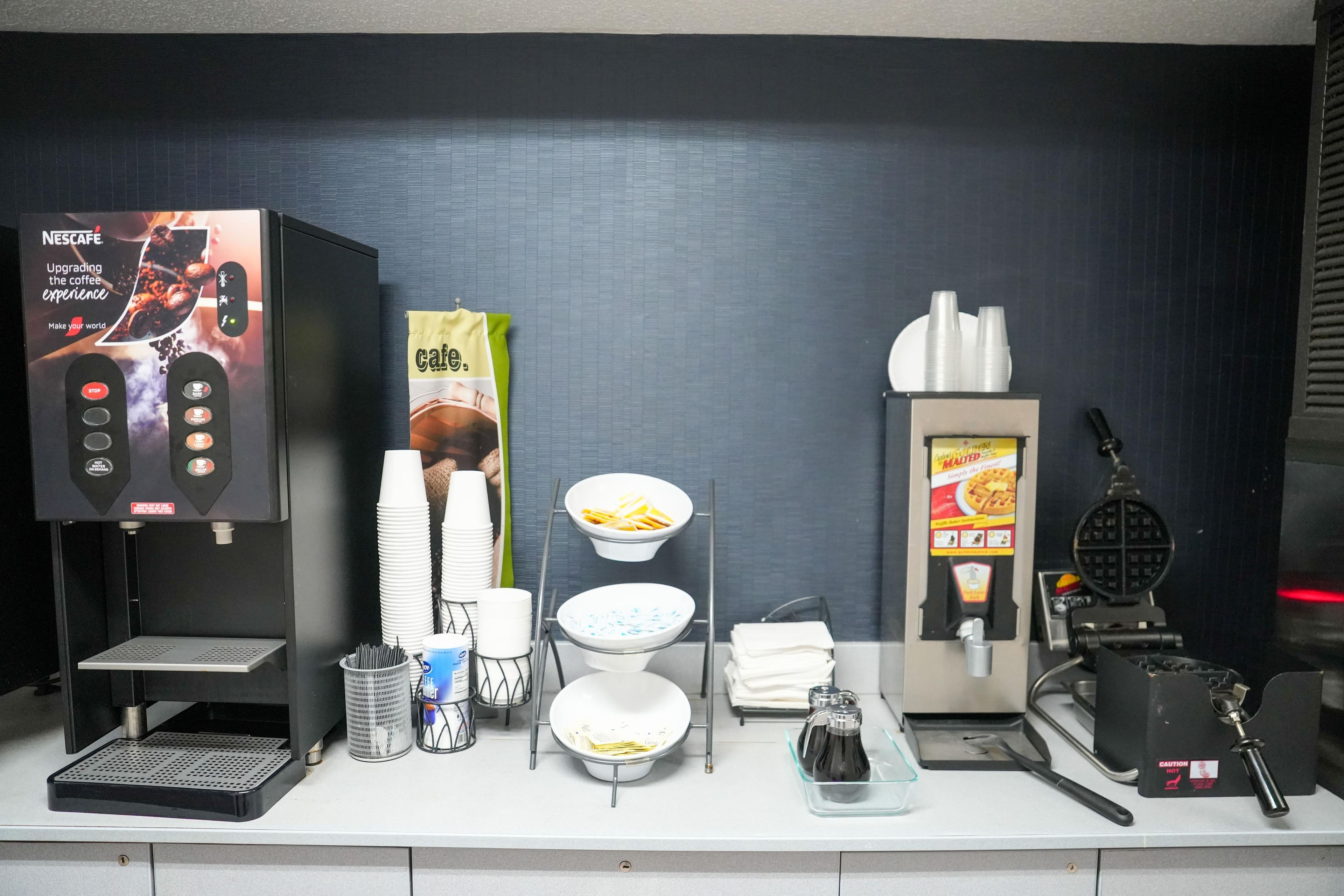 Una estación de café con máquina de Nescafé, plancha para gofres y varias tazas y cuencos.
