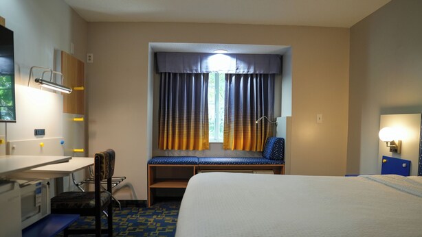 A hotel room with a bed, a desk, a chair, a window with curtains, and a wall-mounted lamp.