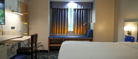 A hotel room with a bed, a desk, a chair, a window with curtains, and a wall-mounted lamp.