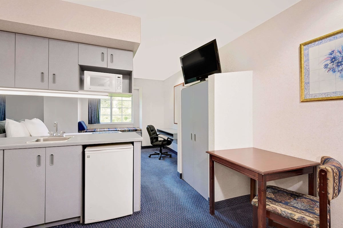 Habitación de hotel compacta con kitchenette, televisor de pantalla plana en la pared, una mesa de comedor con una silla y una cama individual al fondo.