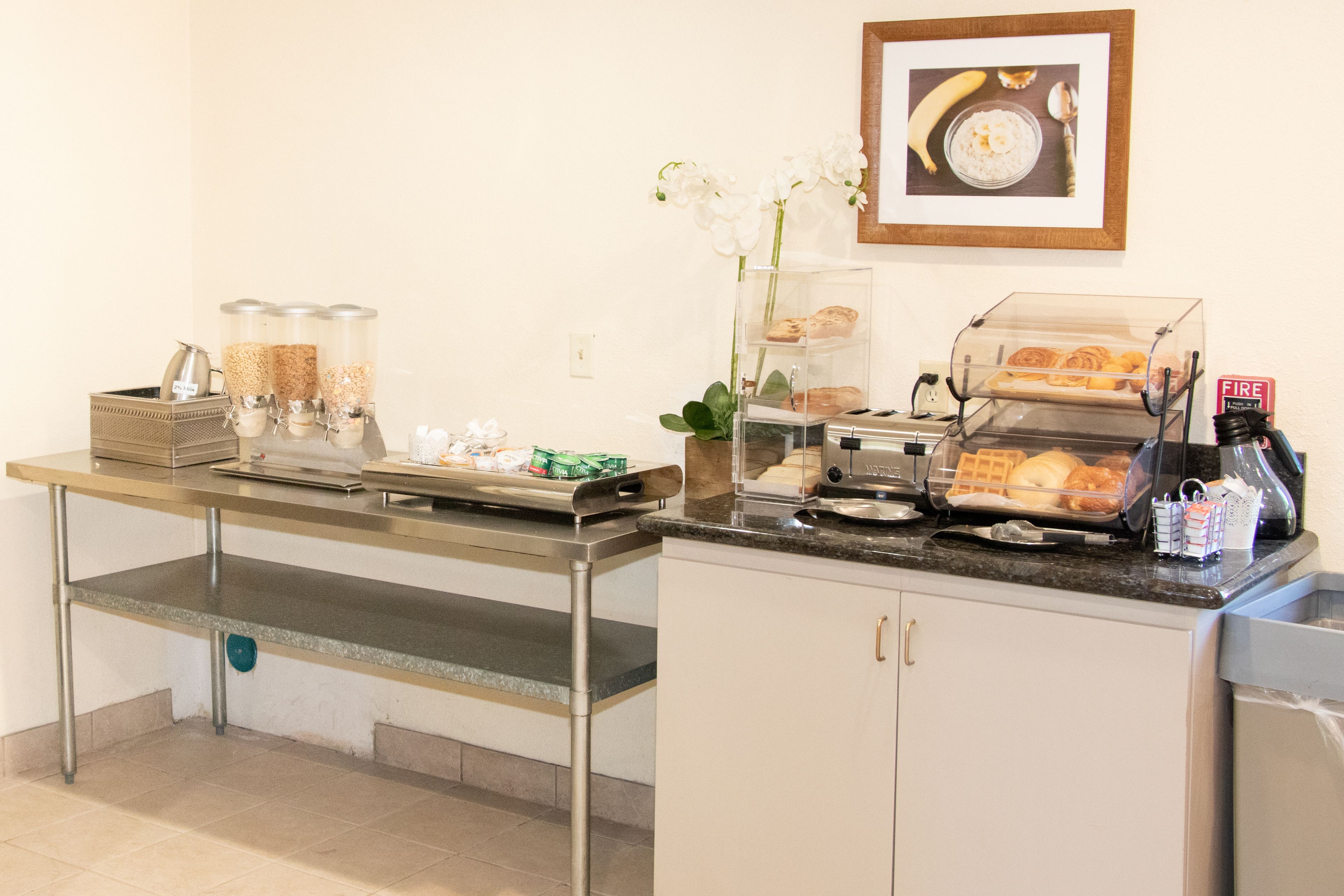 A breakfast bar with coffee machines, a fruit display, and a framed picture on the wall.