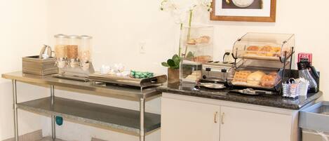 A breakfast bar with coffee machines, a fruit display, and a framed picture on the wall.