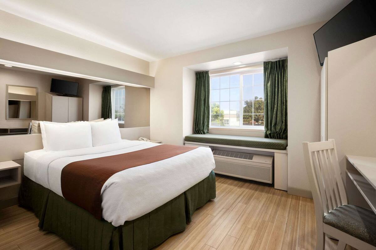 A hotel room with a bed, a desk, a chair, a TV, a window with curtains, and a wall-mounted TV.