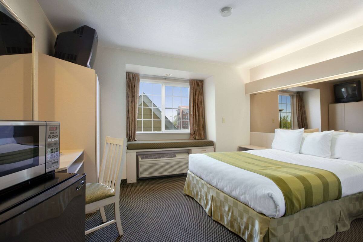 A hotel room with a bed, a desk with a microwave, a chair, a window with curtains, and a television on the wall.