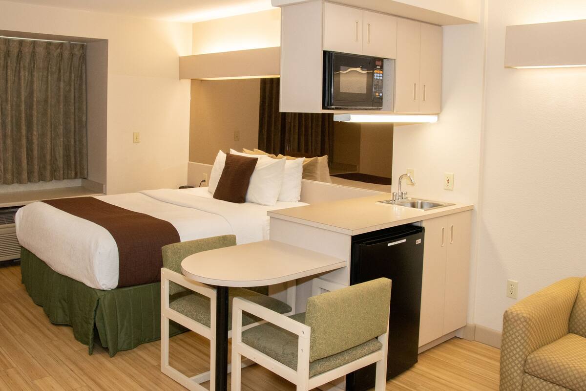 A hotel room with a bed, a small kitchenette, a microwave, a sink, and a dining area with a table and chairs.