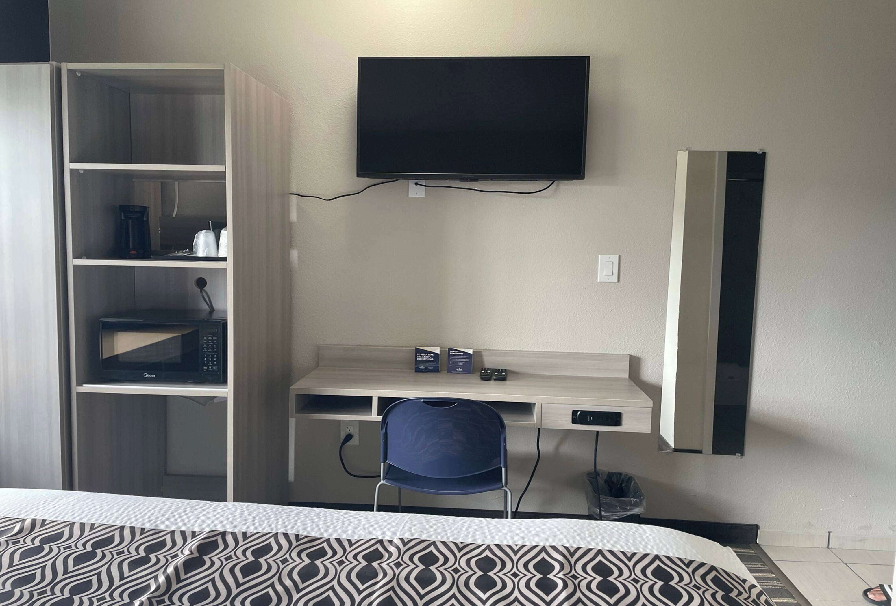 A hotel room with a bed, a desk with a chair, a television mounted on the wall, a wardrobe, and a microwave.