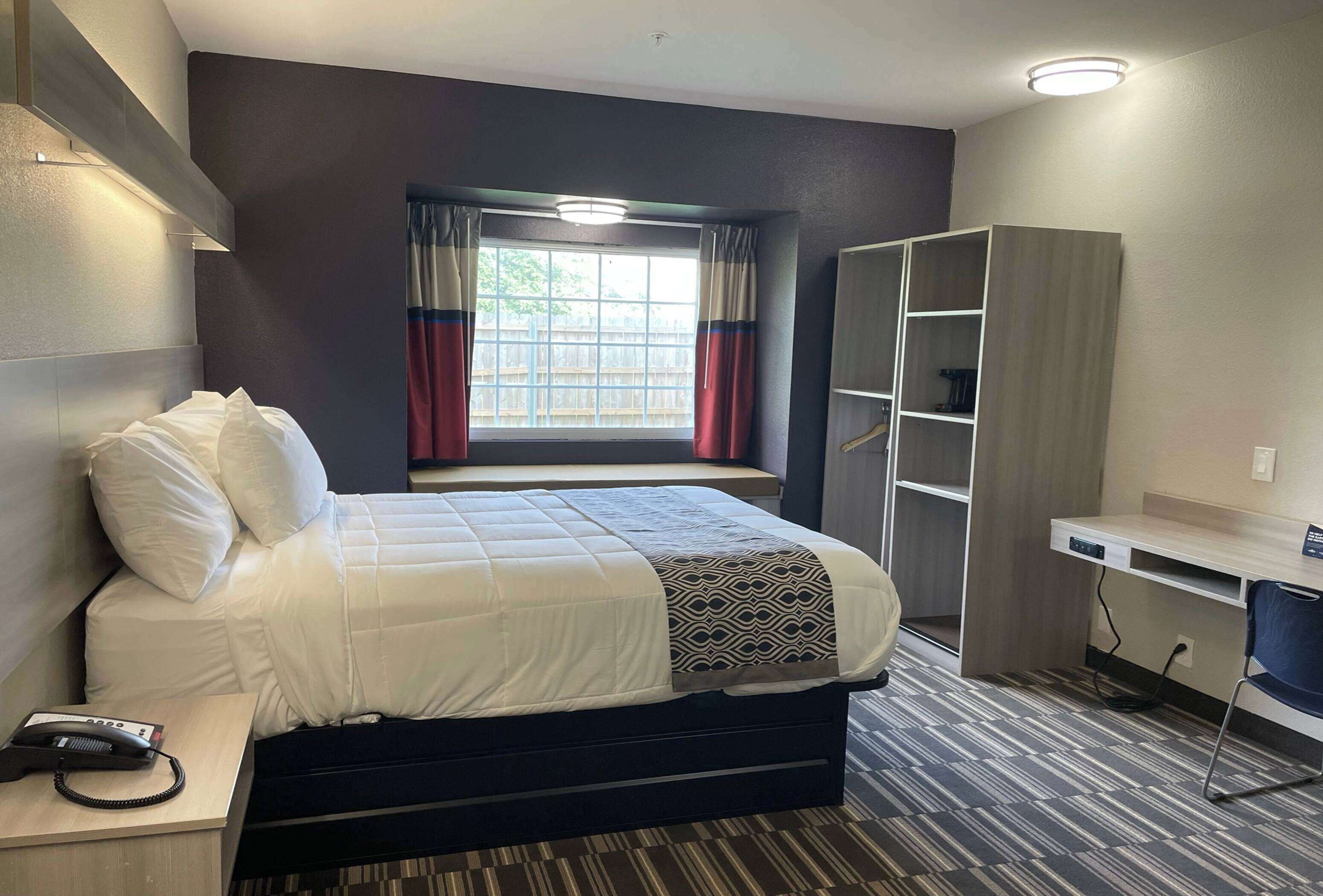 Ein Hotelzimmer mit Bett, Nachttisch mit Telefon, Schreibtisch mit Stuhl, Kleiderschrank und Fenster mit Vorhängen.