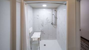 Ducha a pie con pared de azulejos blancos, cabezal de ducha, ducha de mano y una toalla blanca sobre un banco.